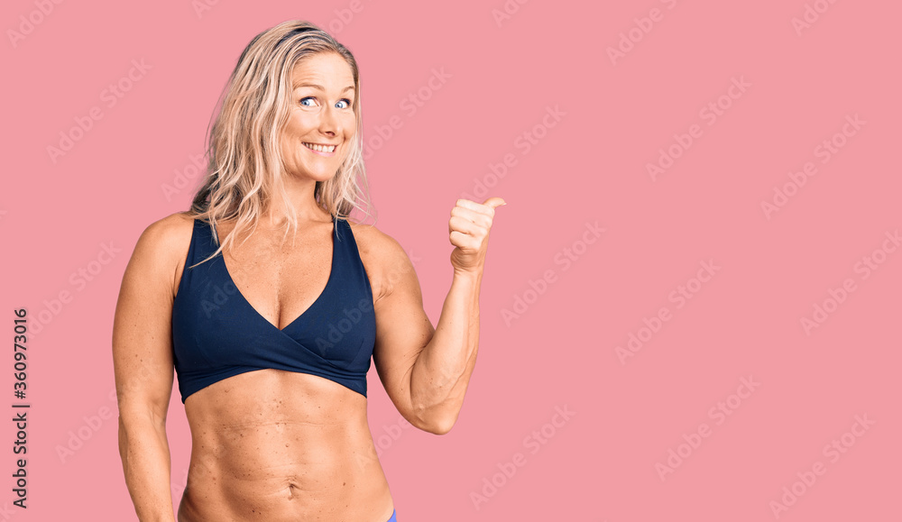 Middle age fit blonde woman wearing bikini smiling with happy face looking and pointing to the side with thumb up. Middle age fit blonde woman wearing bikini smiling with happy face looking and pointing to the side with thumb up.