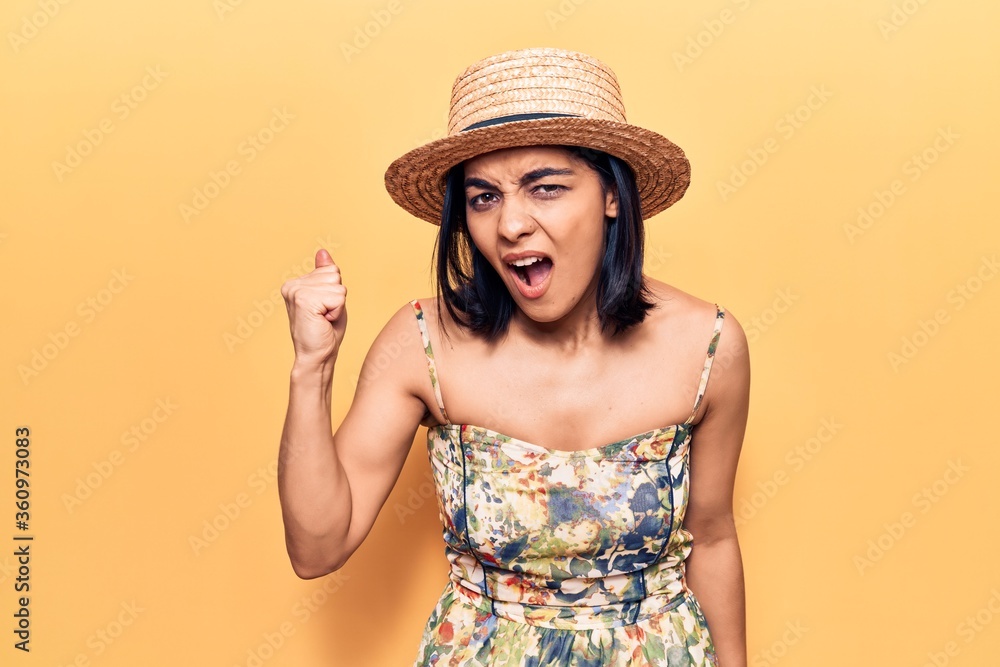 Young beautiful latin woman wearing summer hat angry and mad raising fist frustrated and furious while shouting with anger. rage and aggressive concept. Young beautiful latin woman wearing summer hat angry and mad raising fist frustrated and furious while shouting with anger. rage and aggressive concept.