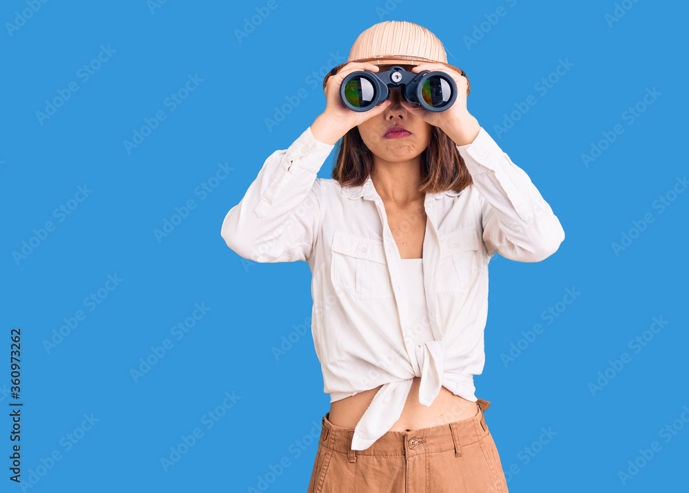 Young beautiful chinese girl wearing explorer hat holding binoculars thinking attitude and sober expression looking self confident Young beautiful chinese girl wearing explorer hat holding binoculars thinking attitude and sober expression looking self confident