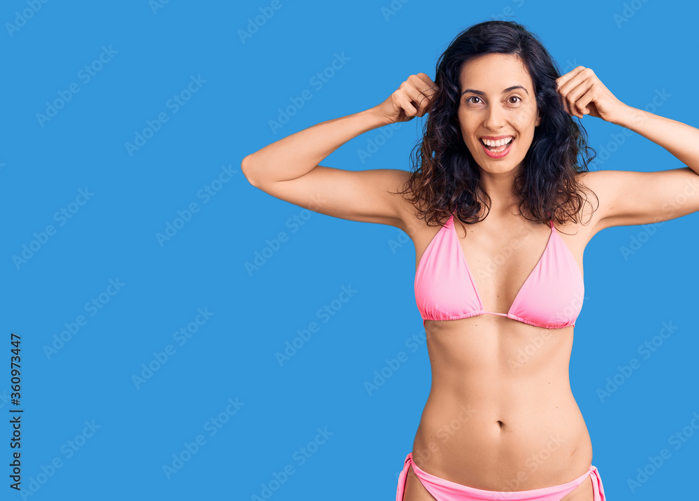 Naklejka premium Young beautiful hispanic woman wearing bikini smiling pulling ears with fingers, funny gesture. audition problem