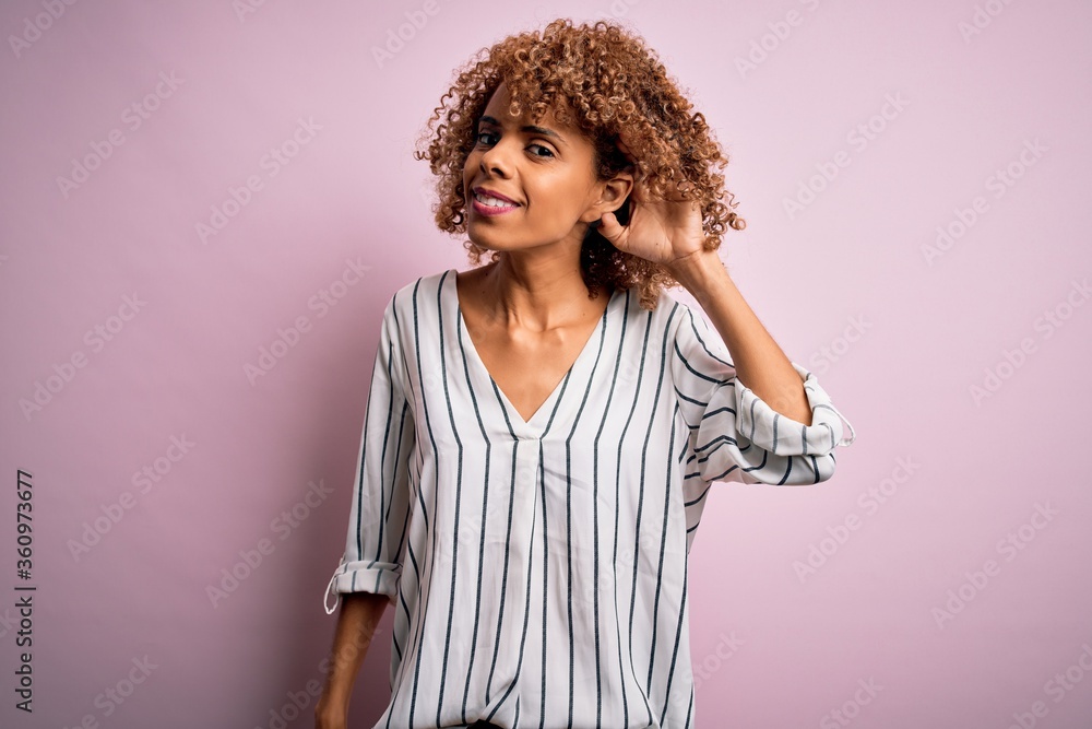 Beautiful african american woman with curly hair wearing striped t-shirt over pink background smiling with hand over ear listening an hearing to rumor or gossip. Deafness concept. Beautiful african american woman with curly hair wearing striped t-shirt over pink background smiling with hand over ear listening an hearing to rumor or gossip. Deafness concept.