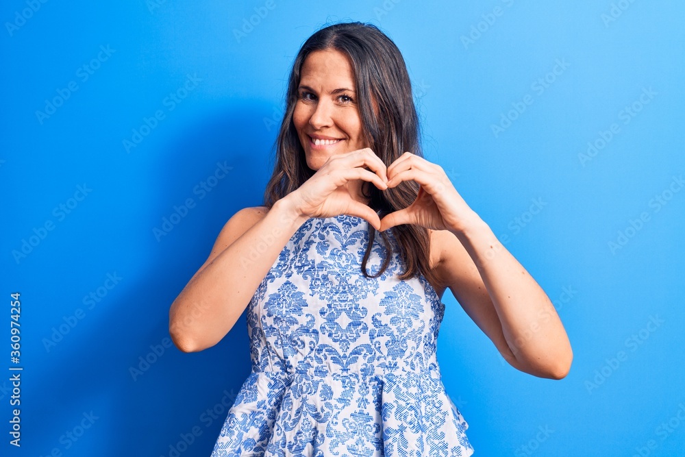 Young beautiful brunette woman wearing casual sleeveless t-shirt over blue background smiling in love doing heart symbol shape with hands. Romantic concept. Young beautiful brunette woman wearing casual sleeveless t-shirt over blue background smiling in love doing heart symbol shape with hands. Romantic concept.
