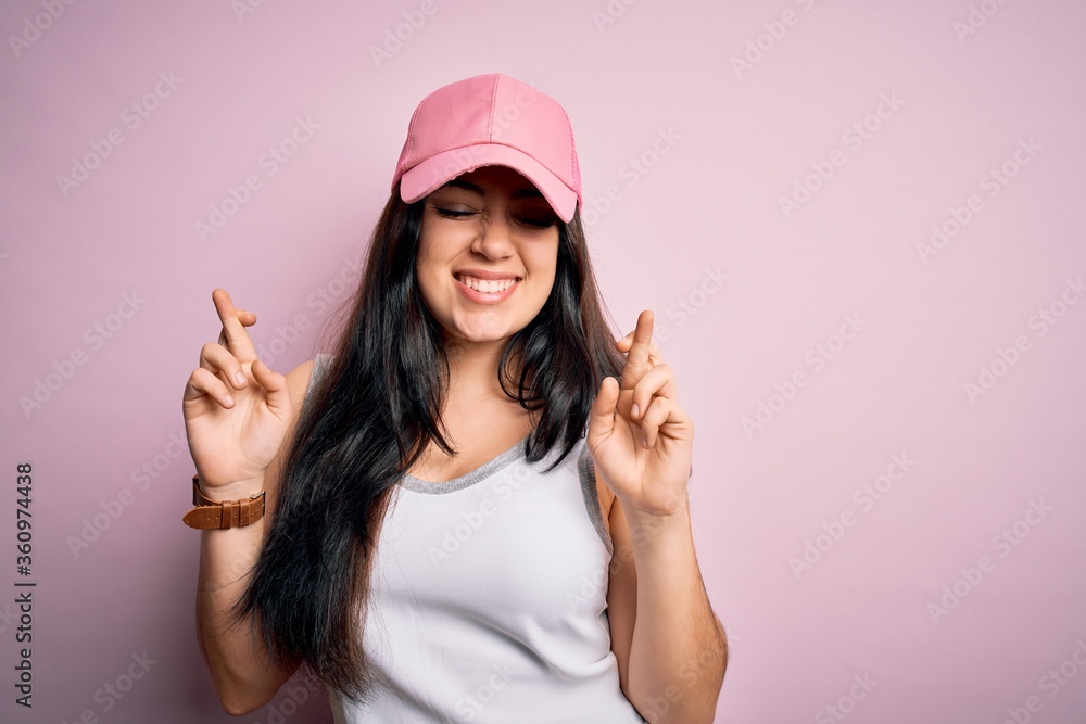 Young brunette woman wearing casual sport cap over pink background gesturing finger crossed smiling with hope and eyes closed. Luck and superstitious concept. Young brunette woman wearing casual sport cap over pink background gesturing finger crossed smiling with hope and eyes closed. Luck and superstitious concept.