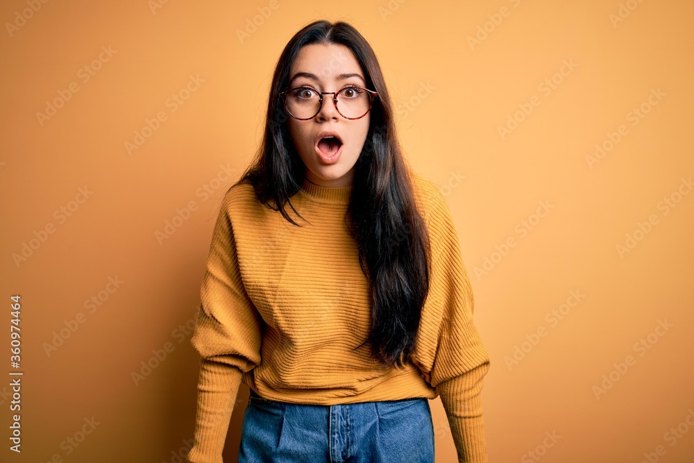 Young brunette woman wearing glasses and casual sweater over yellow isolated background afraid and shocked with surprise and amazed expression, fear and excited face. Young brunette woman wearing glasses and casual sweater over yellow isolated background afraid and shocked with surprise and amazed expression, fear and excited face.