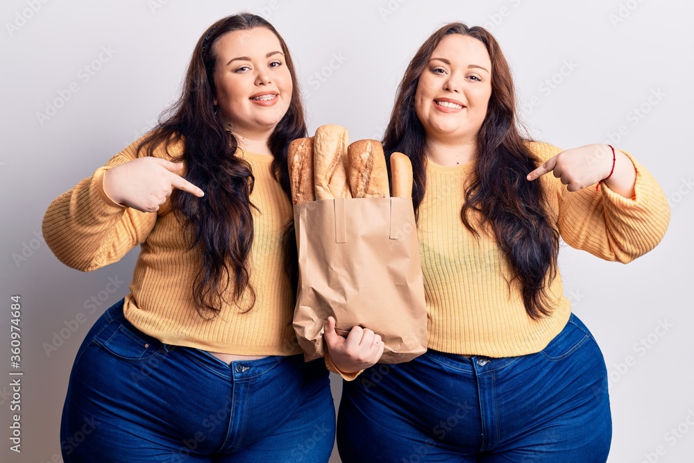Young plus size twins holding paper bag with bread pointing finger to one self smiling happy and proud Young plus size twins holding paper bag with bread pointing finger to one self smiling happy and proud