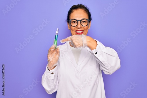 Middle age senior scientist woman wearing laboratory coat holding research test tube very happy pointing with hand and finger