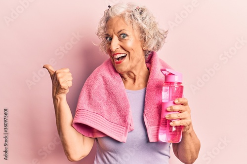 Senior grey-haired woman wearing sportswear and towel drinking bottle of water pointing thumb up to the side smiling happy with open mouth