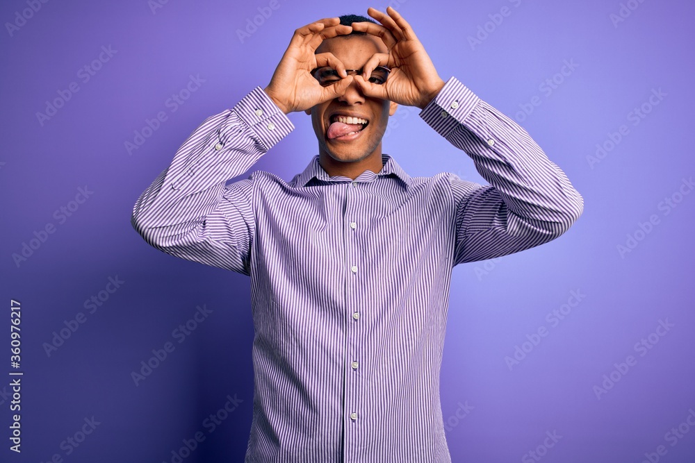 Handsome african american man wearing striped shirt and glasses over purple background doing ok gesture like binoculars sticking tongue out, eyes looking through fingers. Crazy expression.