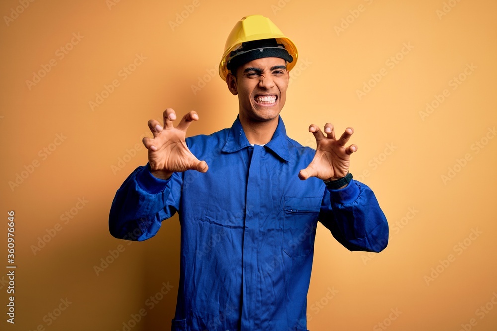 Young handsome african american worker man wearing blue uniform and security helmet smiling funny doing claw gesture as cat, aggressive and sexy expression