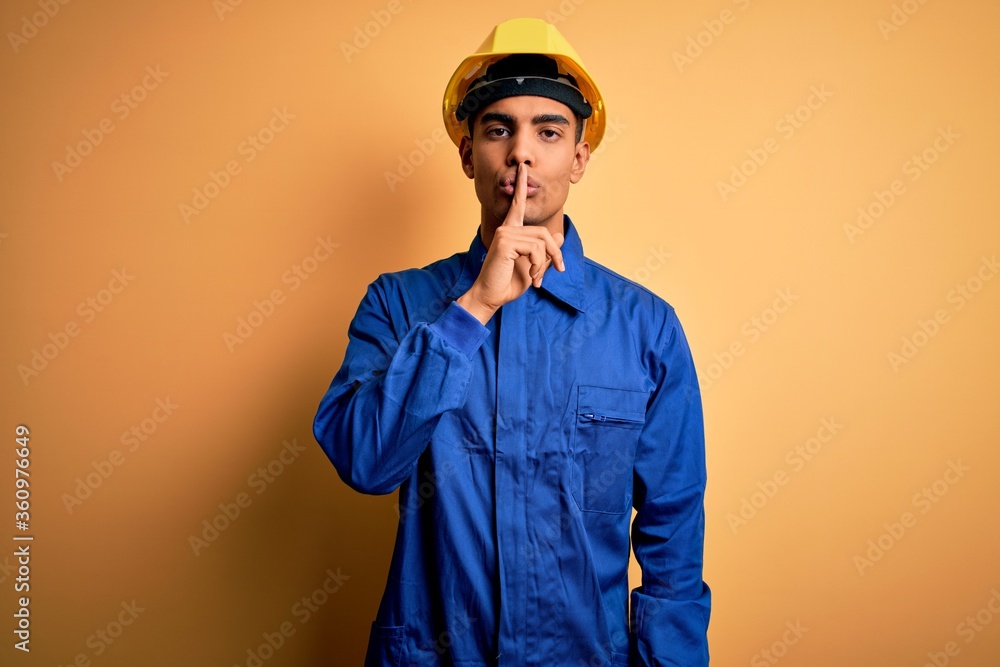 Young handsome african american worker man wearing blue uniform and security helmet asking to be quiet with finger on lips. Silence and secret concept. Young handsome african american worker man wearing blue uniform and security helmet asking to be quiet with finger on lips. Silence and secret concept.
