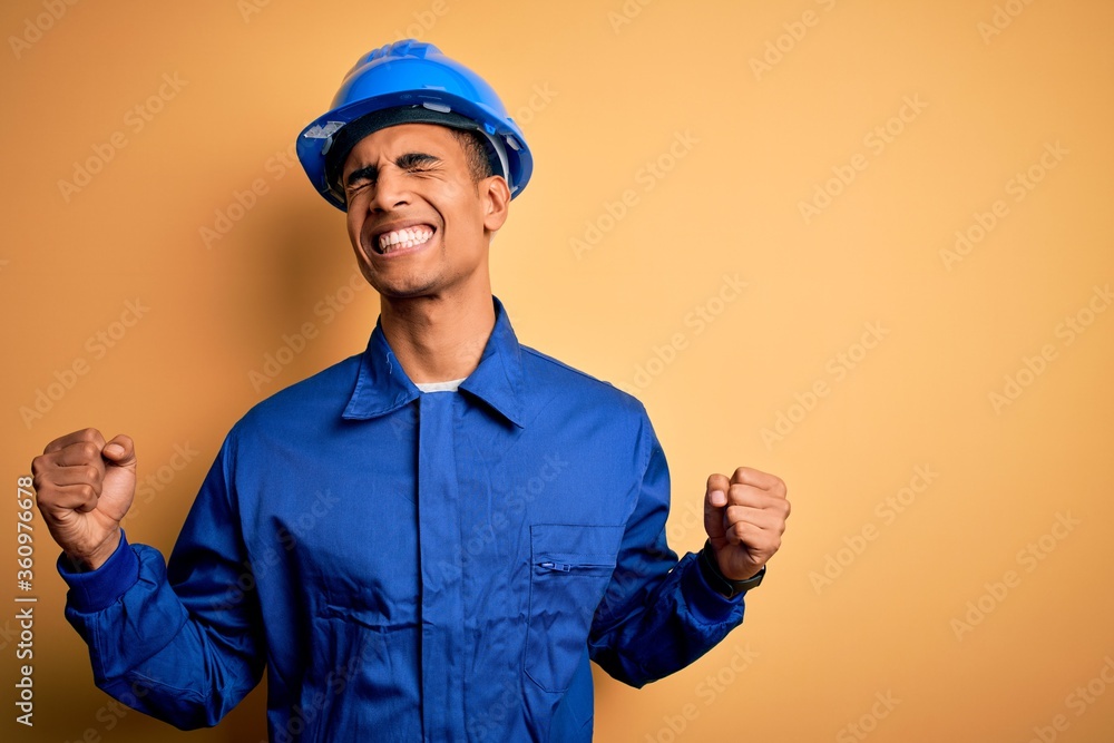 Young handsome african american worker man wearing blue uniform and security helmet very happy and excited doing winner gesture with arms raised, smiling and screaming for success. Celebration. Young handsome african american worker man wearing blue uniform and security helmet very happy and excited doing winner gesture with arms raised, smiling and screaming for success. Celebration.