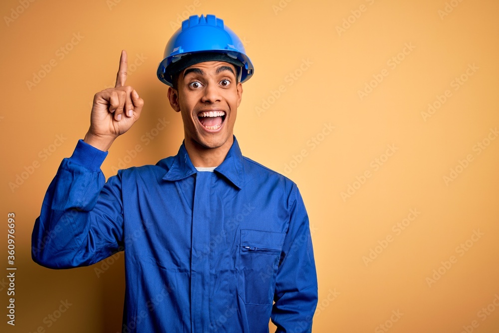 Young handsome african american worker man wearing blue uniform and security helmet pointing finger up with successful idea. Exited and happy. Number one.