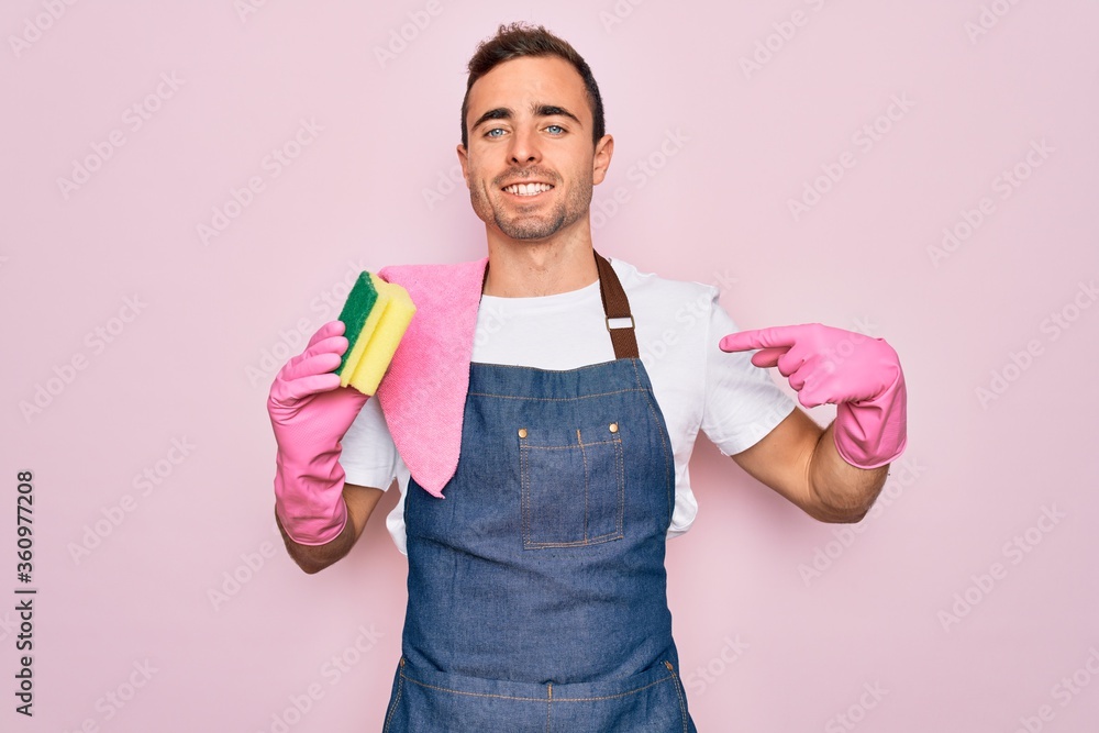 Young cleaner man with blue eyes cleaning wearing apron and gloves using clean scourer with surprise face pointing finger to himself