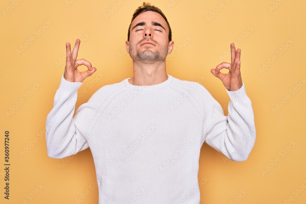 Young handsome man with blue eyes wearing casual sweater standing over yellow background relaxed and smiling with eyes closed doing meditation gesture with fingers. Yoga concept. Young handsome man with blue eyes wearing casual sweater standing over yellow background relaxed and smiling with eyes closed doing meditation gesture with fingers. Yoga concept.