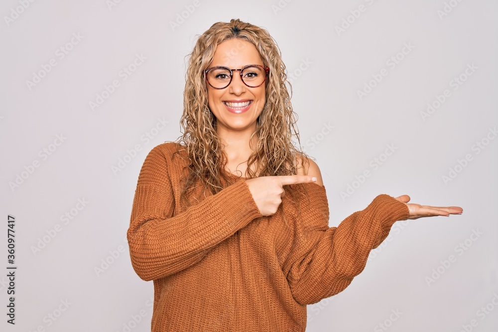 Young beautiful blonde woman wearing casual sweater and glasses over white background amazed and smiling to the camera while presenting with hand and pointing with finger.