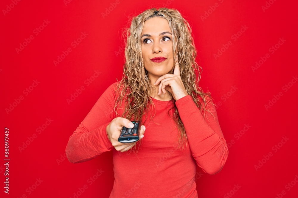 Young beautiful blonde woman using television remote control over isolated red background serious face thinking about question with hand on chin, thoughtful about confusing idea Young beautiful blonde woman using television remote control over isolated red background serious face thinking about question with hand on chin, thoughtful about confusing idea
