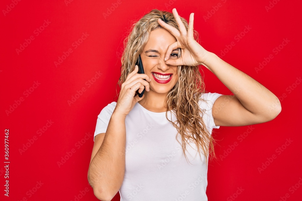 Beautiful blonde woman having conversation talking on the smartphone over red background smiling happy doing ok sign with hand on eye looking through fingers