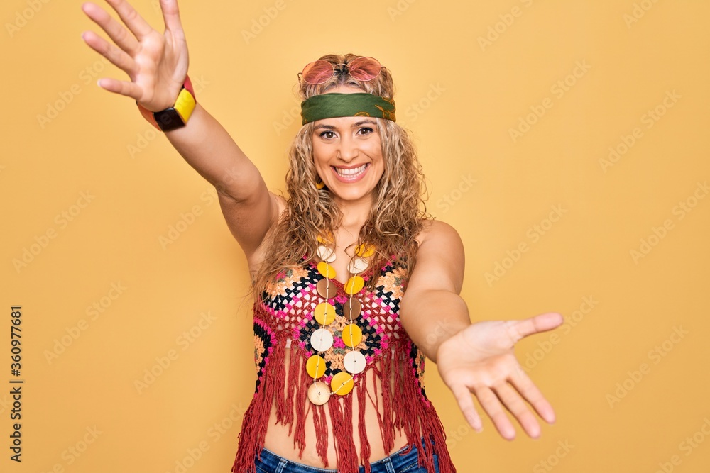 Beautiful blonde hippie woman wearing sunglasses accessories over yellow background looking at the camera smiling with open arms for hug. Cheerful expression embracing happiness. Photo | Adobe Stock