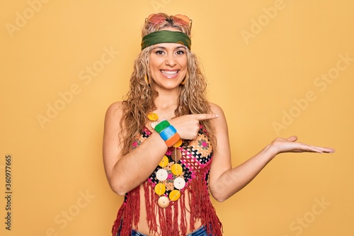 Beautiful blonde hippie woman wearing sunglasses and accessories over yellow background amazed and smiling to the camera while presenting with hand and pointing with finger.