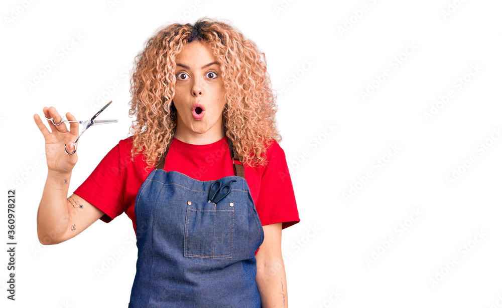 Young blonde woman with curly hair wearing barber apron scared and amazed with open mouth for surprise, disbelief face Young blonde woman with curly hair wearing barber apron scared and amazed with open mouth for surprise, disbelief face