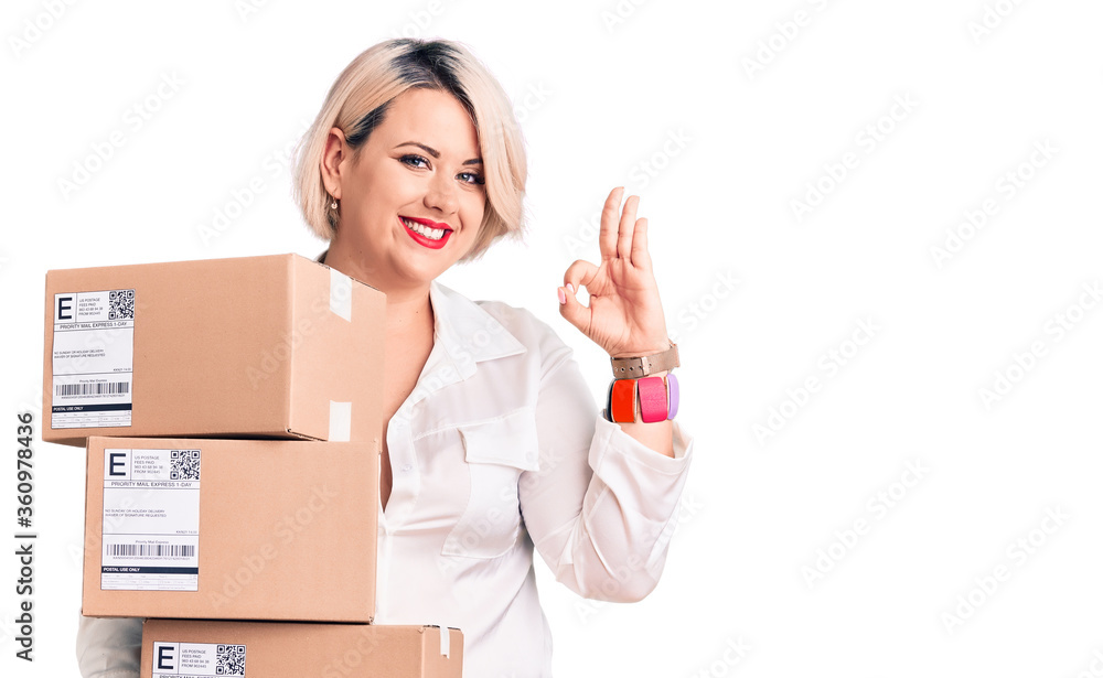 Young blonde plus size woman holding delivery cardboard package doing ok sign with fingers, smiling friendly gesturing excellent symbol Young blonde plus size woman holding delivery cardboard package doing ok sign with fingers, smiling friendly gesturing excellent symbol