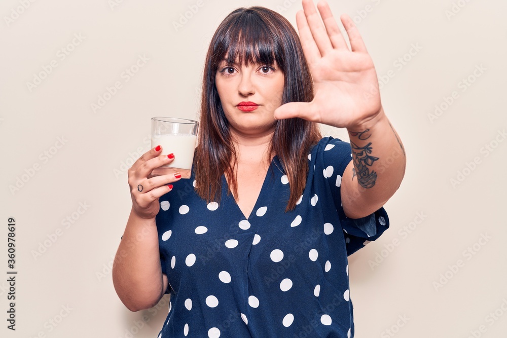 Young plus size woman holding glass of milk with open hand doing stop sign with serious and confident expression, defense gesture Young plus size woman holding glass of milk with open hand doing stop sign with serious and confident expression, defense gesture
