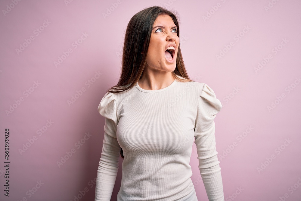 Young beautiful woman with blue eyes wearing casual white t-shirt over pink background angry and mad screaming frustrated and furious, shouting with anger. Rage and aggressive concept. Young beautiful woman with blue eyes wearing casual white t-shirt over pink background angry and mad screaming frustrated and furious, shouting with anger. Rage and aggressive concept.