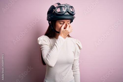 Young beautiful motorcyclist woman with blue eyes wearing moto helmet over pink background tired rubbing nose and eyes feeling fatigue and headache. Stress and frustration concept. © Krakenimages.com