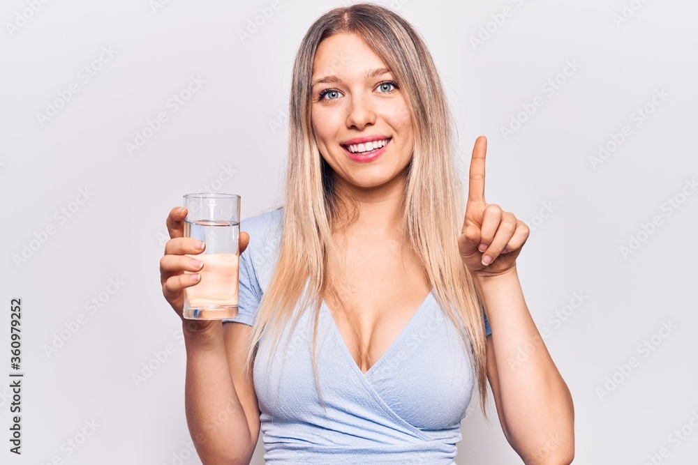 Young beautiful blonde girl drinking glass of water smiling with an idea or question pointing finger with happy face, number one Young beautiful blonde girl drinking glass of water smiling with an idea or question pointing finger with happy face, number one