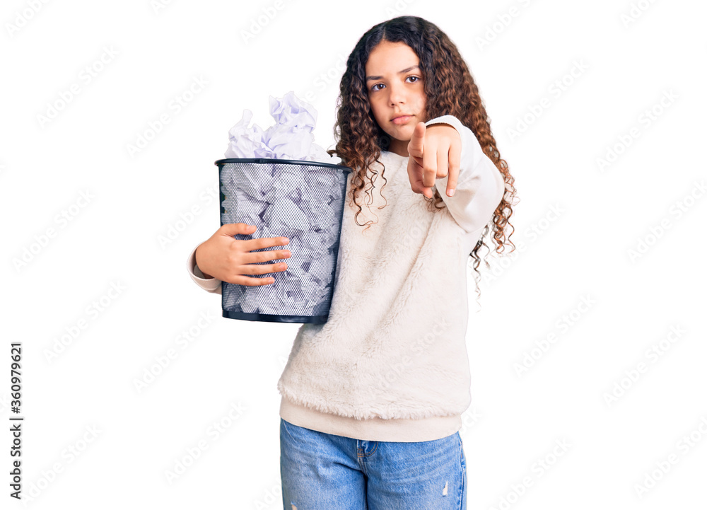 Beautiful kid girl with curly hair holding paper bin full of crumpled papers pointing with finger to the camera and to you, confident gesture looking serious Beautiful kid girl with curly hair holding paper bin full of crumpled papers pointing with finger to the camera and to you, confident gesture looking serious