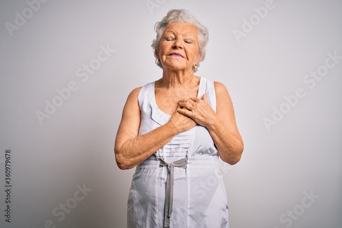 Senior beautiful grey-haired woman wearing casual summer dress over white background smiling with hands on chest with closed eyes and grateful gesture on face. Health concept.