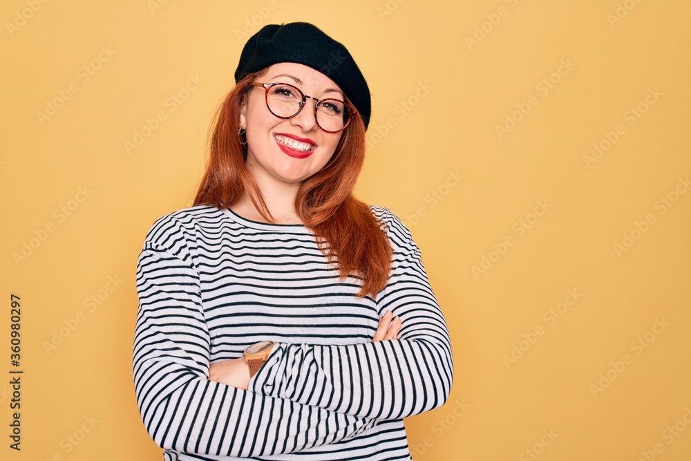 Beautiful redhead woman wearing striped t-shirt and french beret over yellow background happy face smiling with crossed arms looking at the camera. Positive person.