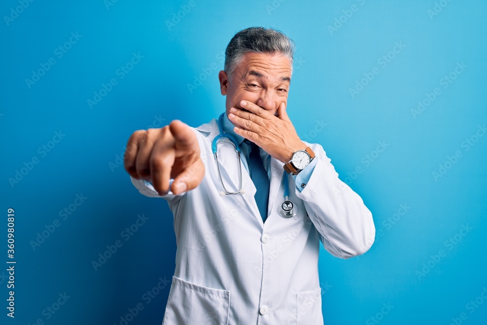 Middle age handsome grey-haired doctor man wearing coat and blue stethoscope laughing at you, pointing finger to the camera with hand over mouth, shame expression Middle age handsome grey-haired doctor man wearing coat and blue stethoscope laughing at you, pointing finger to the camera with hand over mouth, shame expression