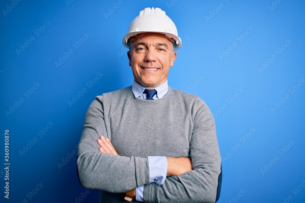 Middle age handsome grey-haired engineer man wearing safety helmet over blue background happy face smiling with crossed arms looking at the camera. Positive person. Middle age handsome grey-haired engineer man wearing safety helmet over blue background happy face smiling with crossed arms looking at the camera. Positive person.
