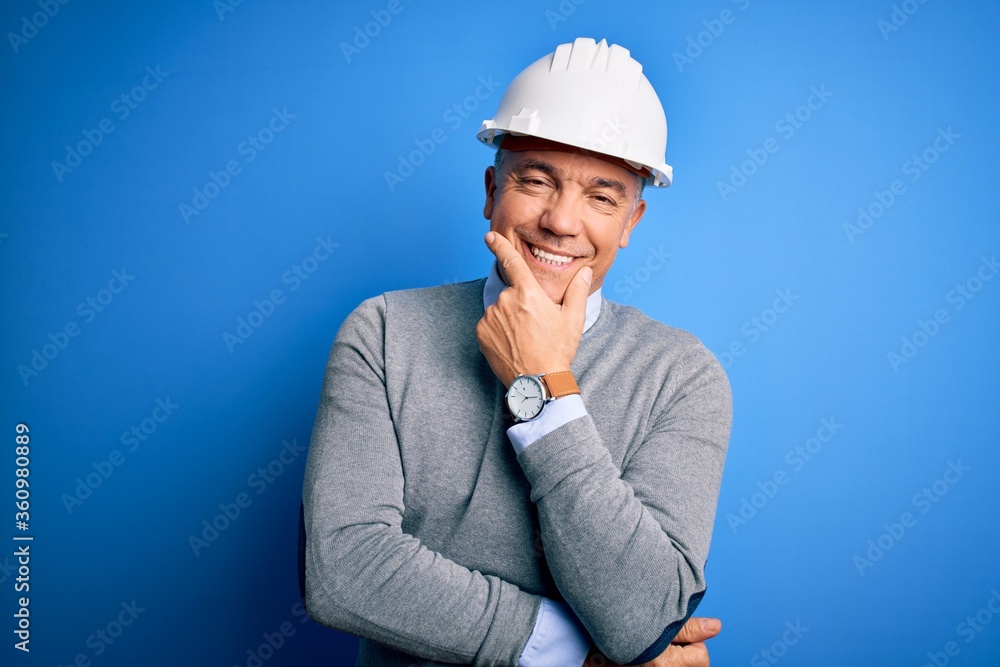 Middle age handsome grey-haired engineer man wearing safety helmet over blue background looking confident at the camera smiling with crossed arms and hand raised on chin. Thinking positive. Middle age handsome grey-haired engineer man wearing safety helmet over blue background looking confident at the camera smiling with crossed arms and hand raised on chin. Thinking positive.