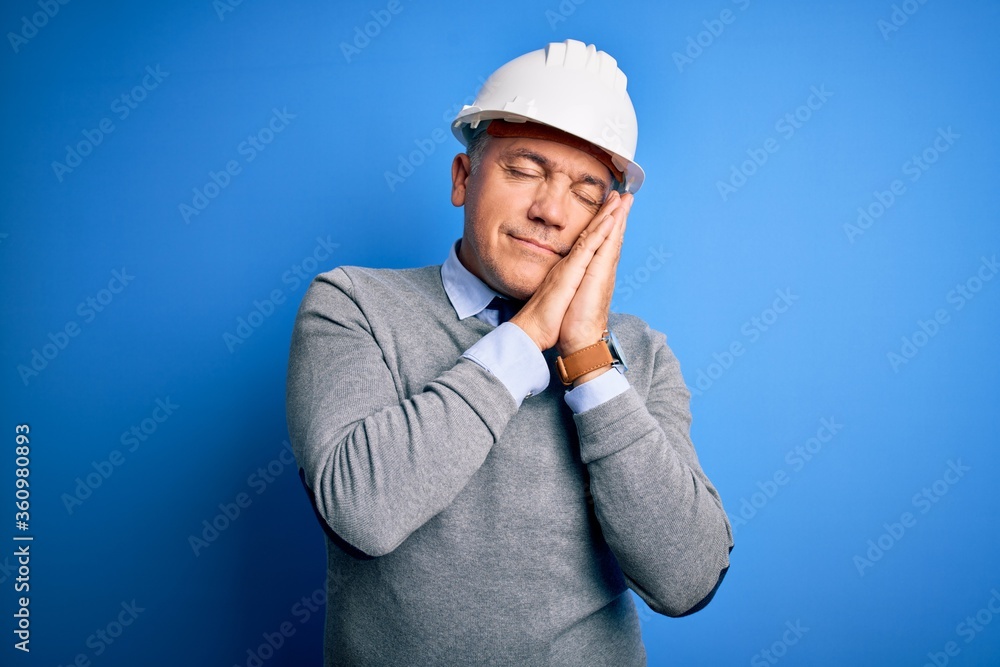 Middle age handsome grey-haired engineer man wearing safety helmet over blue background sleeping tired dreaming and posing with hands together while smiling with closed eyes.