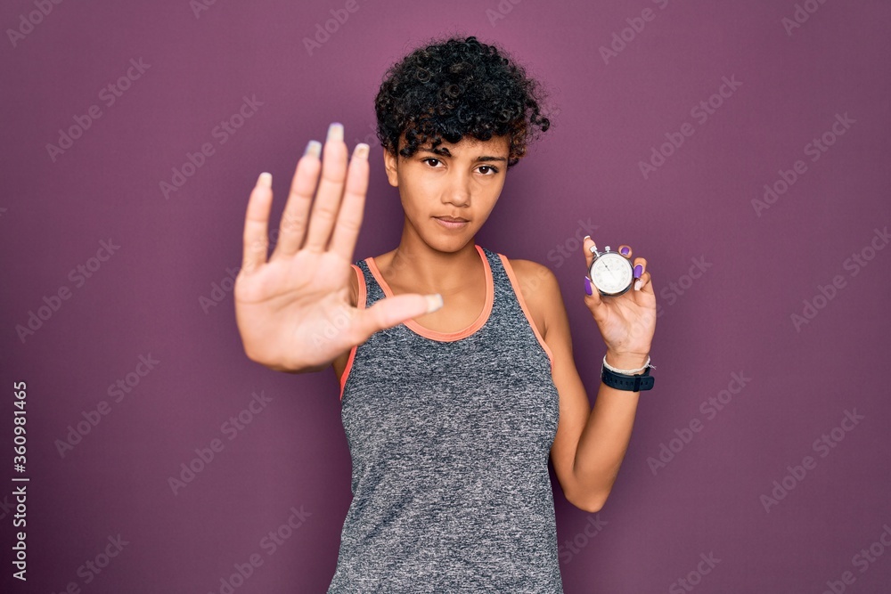 Young beautiful african american sporty woman holding stopwatch over purple background with open hand doing stop sign with serious and confident expression, defense gesture Young beautiful african american sporty woman holding stopwatch over purple background with open hand doing stop sign with serious and confident expression, defense gesture