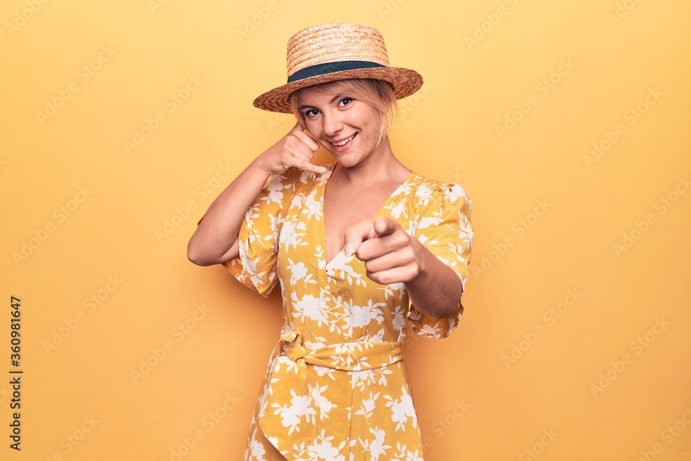 Beautiful blonde woman on vacation wearing summer hat and dress over yellow background smiling doing talking on the telephone gesture and pointing to you. Call me. Beautiful blonde woman on vacation wearing summer hat and dress over yellow background smiling doing talking on the telephone gesture and pointing to you. Call me.
