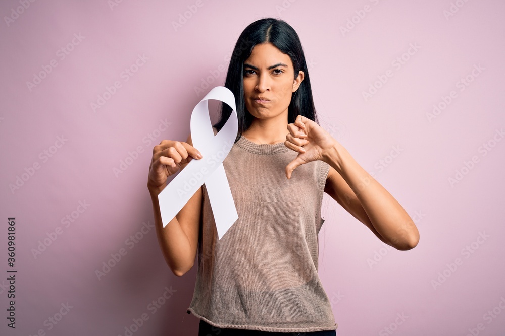 Young beautiful hispanic woman holding white ribbon as stop violence and lung cancer symbol with angry face, negative sign showing dislike with thumbs down, rejection concept Young beautiful hispanic woman holding white ribbon as stop violence and lung cancer symbol with angry face, negative sign showing dislike with thumbs down, rejection concept