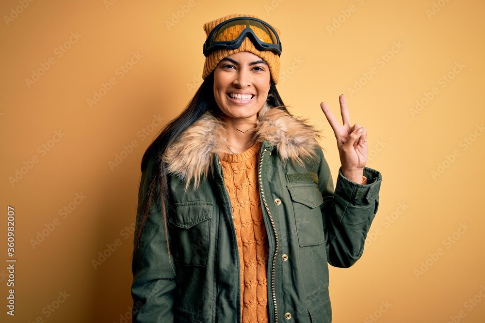 Young beautiful hispanic woman wearing ski glasses and coat for winter weather showing and pointing up with fingers number two while smiling confident and happy. Young beautiful hispanic woman wearing ski glasses and coat for winter weather showing and pointing up with fingers number two while smiling confident and happy.