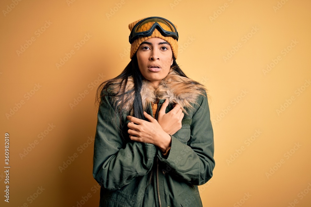 Young beautiful hispanic woman wearing ski glasses and coat for winter weather shaking and freezing for winter cold with sad and shock expression on face Young beautiful hispanic woman wearing ski glasses and coat for winter weather shaking and freezing for winter cold with sad and shock expression on face