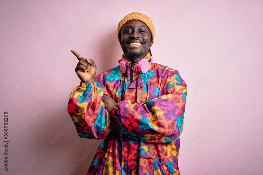Obraz premium Young handsome african american man wearing colorful coat and cap over pink background with a big smile on face, pointing with hand and finger to the side looking at the camera.