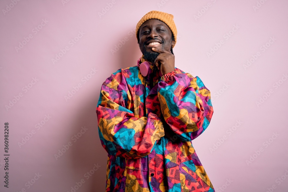 Young handsome african american man wearing colorful coat and cap over pink background looking confident at the camera with smile with crossed arms and hand raised on chin. Thinking positive. Young handsome african american man wearing colorful coat and cap over pink background looking confident at the camera with smile with crossed arms and hand raised on chin. Thinking positive.