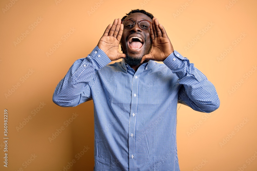 Young handsome african american man wearing shirt and glasses over yellow background Smiling cheerful playing peek a boo with hands showing face. Surprised and exited Young handsome african american man wearing shirt and glasses over yellow background Smiling cheerful playing peek a boo with hands showing face. Surprised and exited