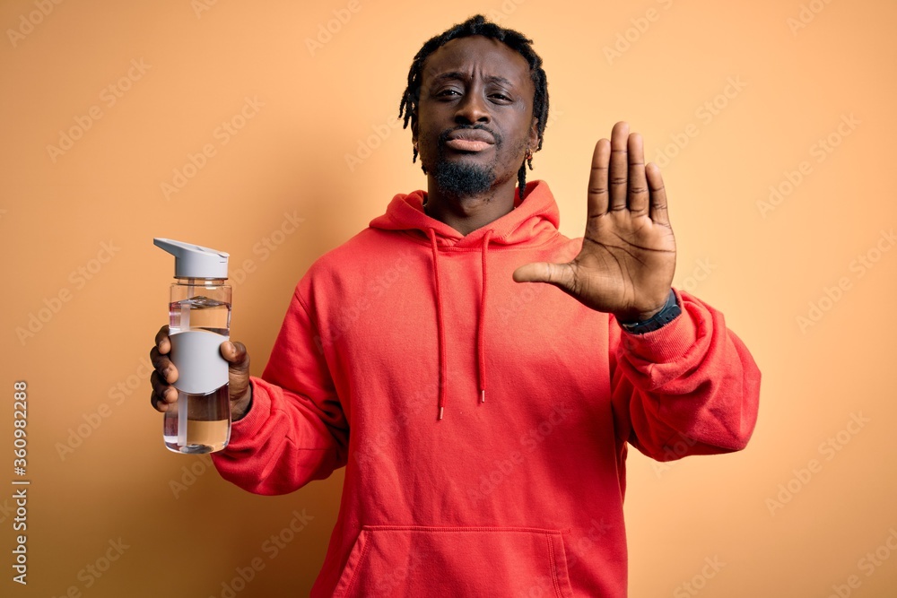 Young african american sporty man wearing sweatshirt with hoodie drinking bottle of water with open hand doing stop sign with serious and confident expression, defense gesture Young african american sporty man wearing sweatshirt with hoodie drinking bottle of water with open hand doing stop sign with serious and confident expression, defense gesture