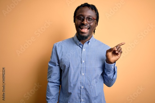 Young handsome african american man wearing shirt and glasses over yellow background with a big smile on face, pointing with hand finger to the side looking at the camera. © Krakenimages.com