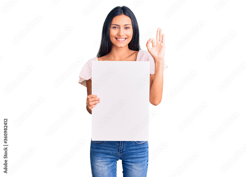Young beautiful latin girl holding blank empty banner doing ok sign with fingers, smiling friendly gesturing excellent symbol Young beautiful latin girl holding blank empty banner doing ok sign with fingers, smiling friendly gesturing excellent symbol