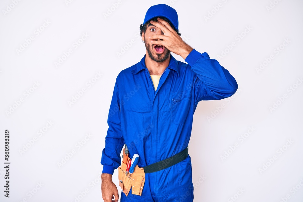 Handsome young man with curly hair and bear weaing handyman uniform peeking in shock covering face and eyes with hand, looking through fingers with embarrassed expression. Handsome young man with curly hair and bear weaing handyman uniform peeking in shock covering face and eyes with hand, looking through fingers with embarrassed expression.