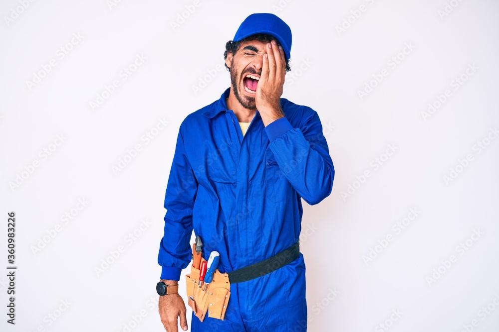 Handsome young man with curly hair and bear weaing handyman uniform yawning tired covering half face, eye and mouth with hand. face hurts in pain. Handsome young man with curly hair and bear weaing handyman uniform yawning tired covering half face, eye and mouth with hand. face hurts in pain.