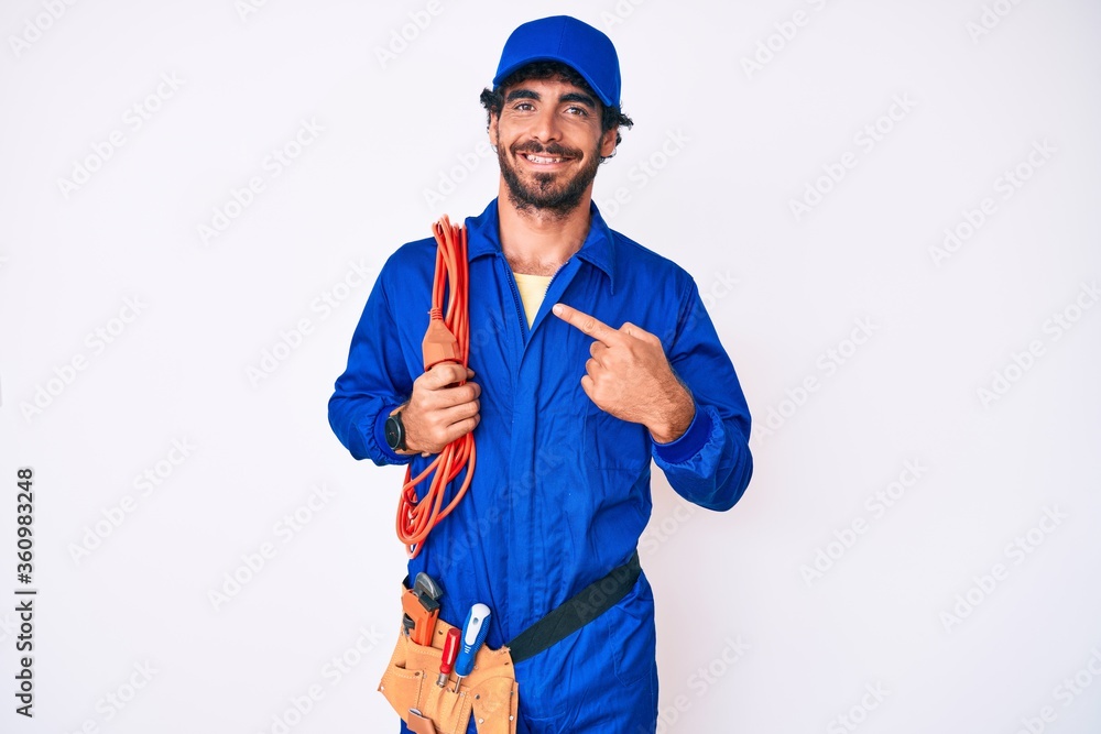 Handsome young man with curly hair and bear wearing builder jumpsuit uniform and electric cables smiling happy pointing with hand and finger Handsome young man with curly hair and bear wearing builder jumpsuit uniform and electric cables smiling happy pointing with hand and finger